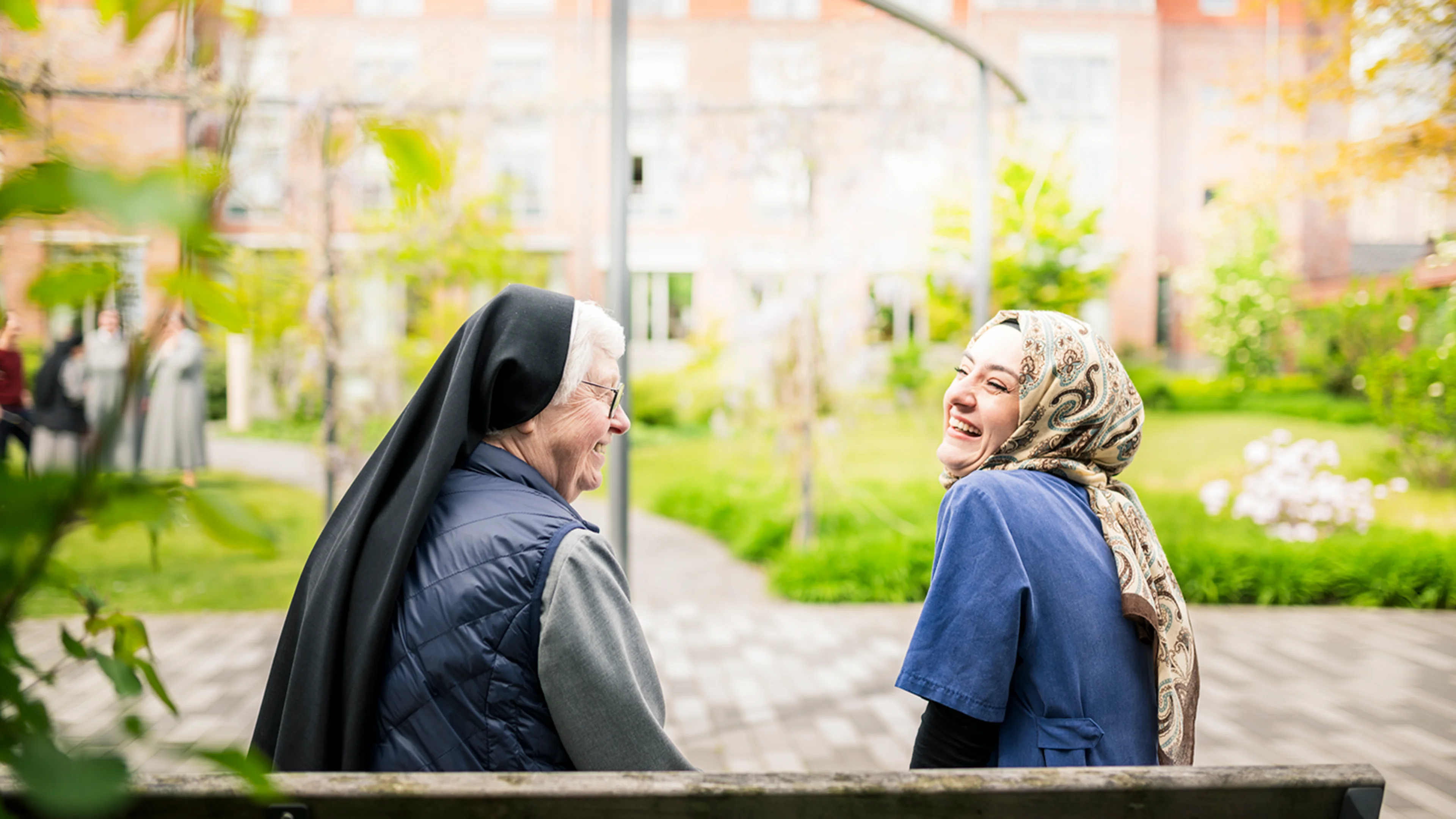 Eine ältere Nonne und eine junge Frau mit Hijab sitzen draußen auf einer Bank und lächeln und lachen gemeinsam in einem hellgrünen Garten, im Hintergrund ist ein Gebäude zu sehen.